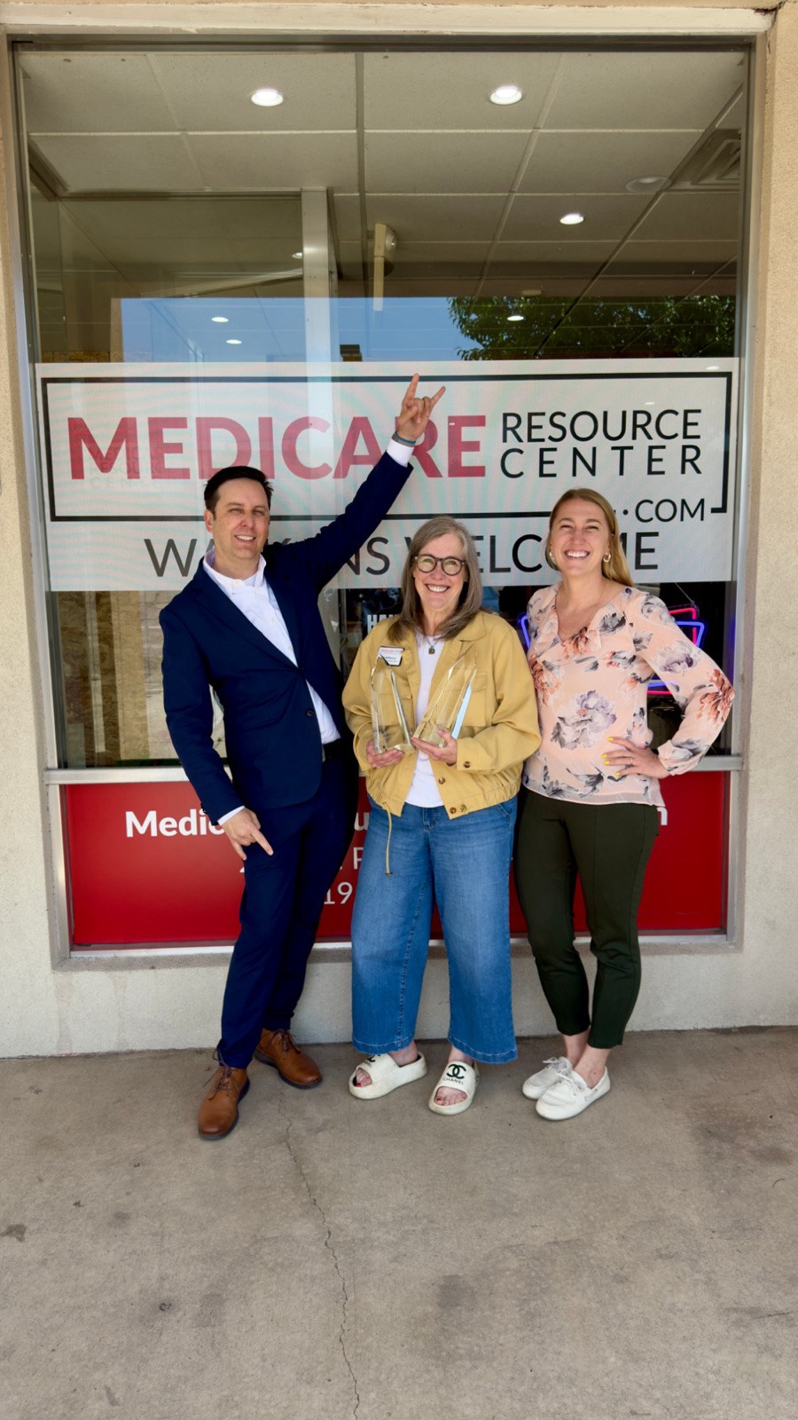 Three Medicare Resource Center agents smiling in front of their Colorado Springs office window, which reads Walk-ins welcome